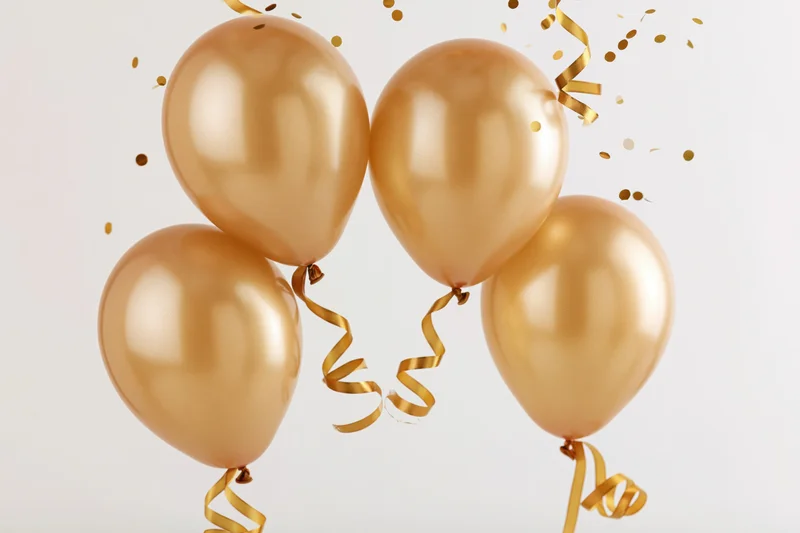 Four gold metallic balloons floating in a loose arch with confetti and ribbons against a white background