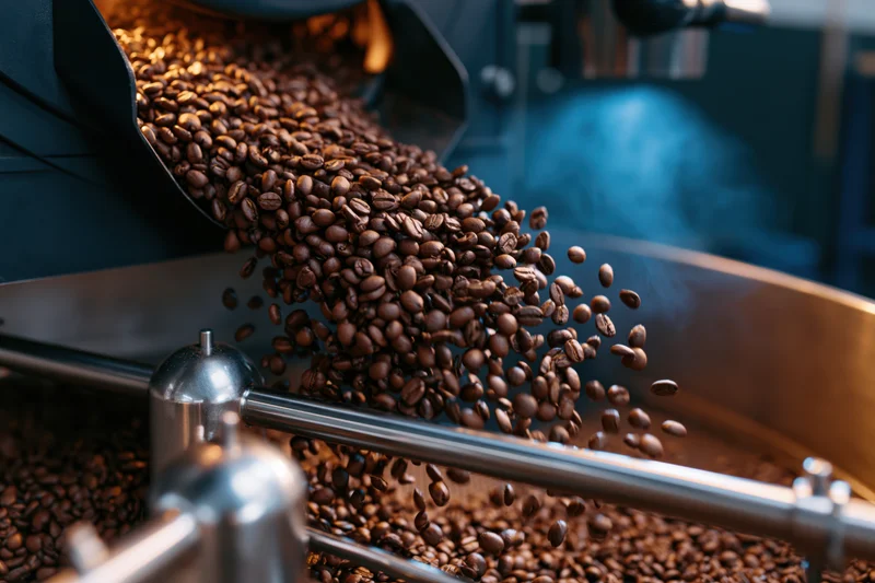 Roasted coffee beans cascading midair from industrial roasting machine chute into metal container