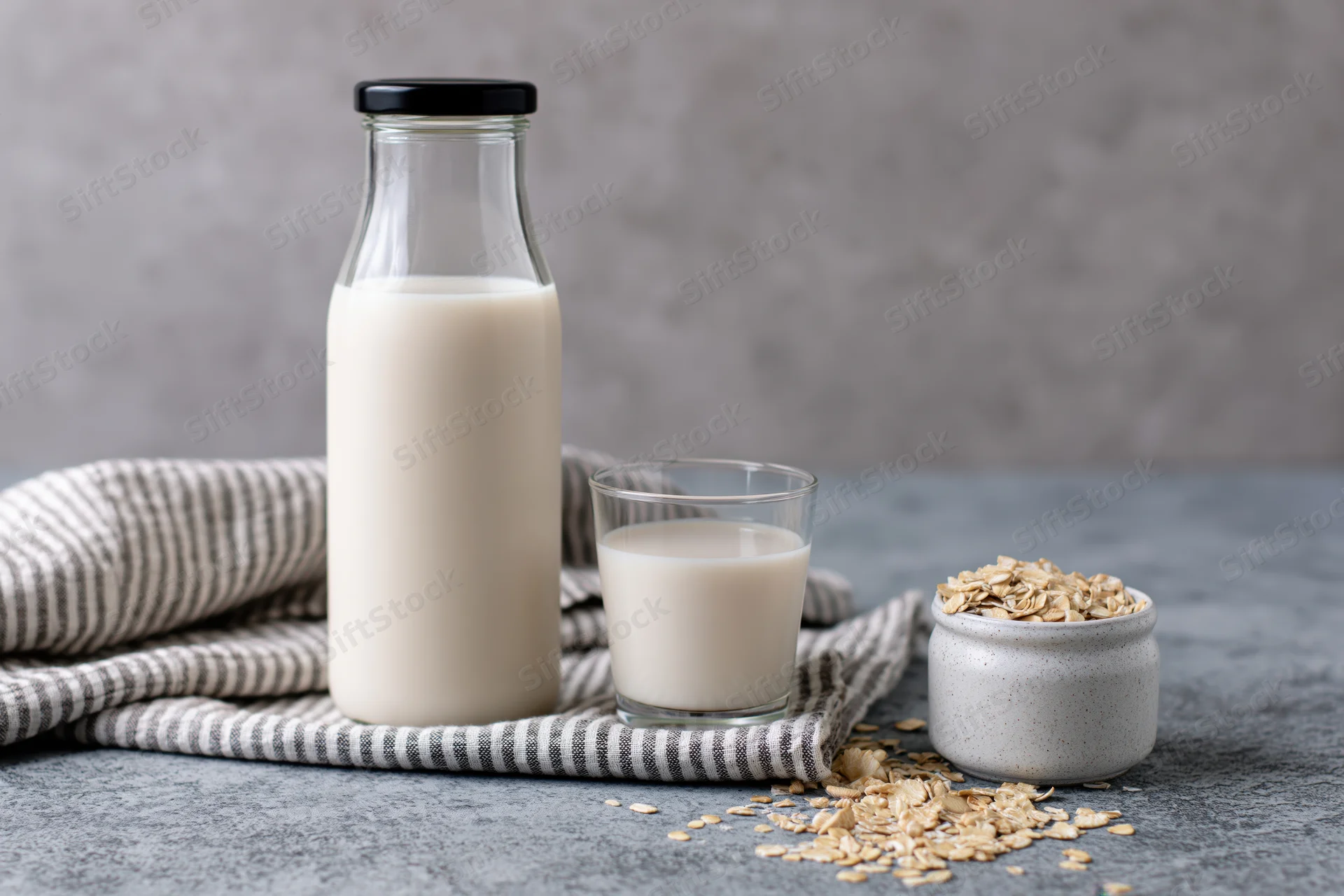Glass bottle of oat milk with black lid and a short drinking glass beside a jar of oats on a striped cloth on a gray countertop