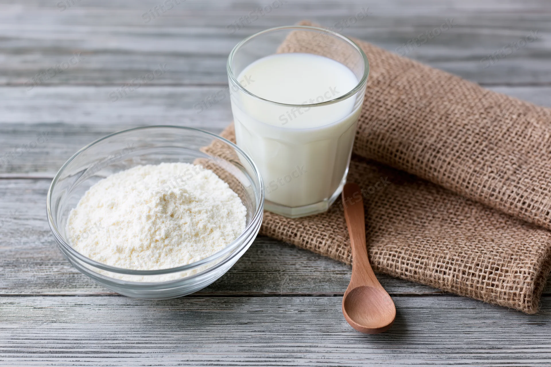 Glass bowl of powdered milk beside a full glass of milk on a wooden table with a wooden spoon and burlap cloth