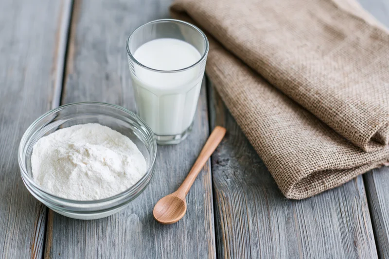 Glass bowl of powdered milk beside a full glass of milk on a wooden table with a wooden spoon and folded burlap cloth