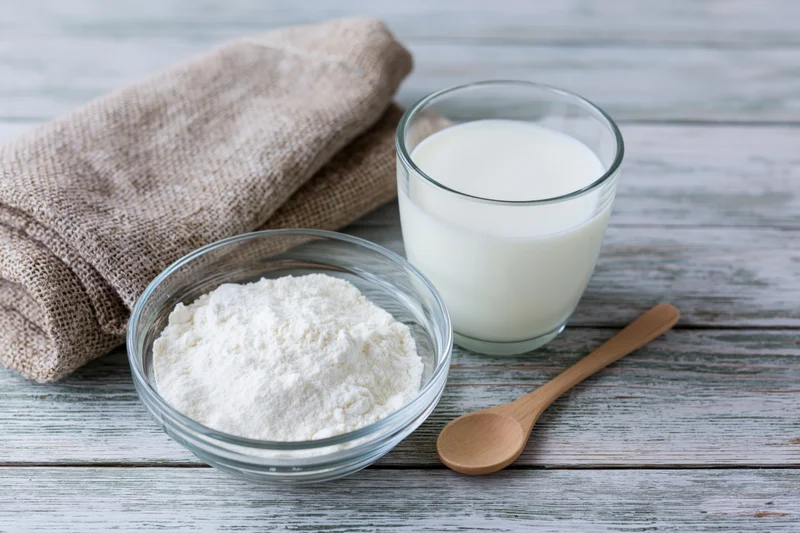 Glass bowl of powdered milk beside a full glass of milk and wooden spoon on a rustic wooden table