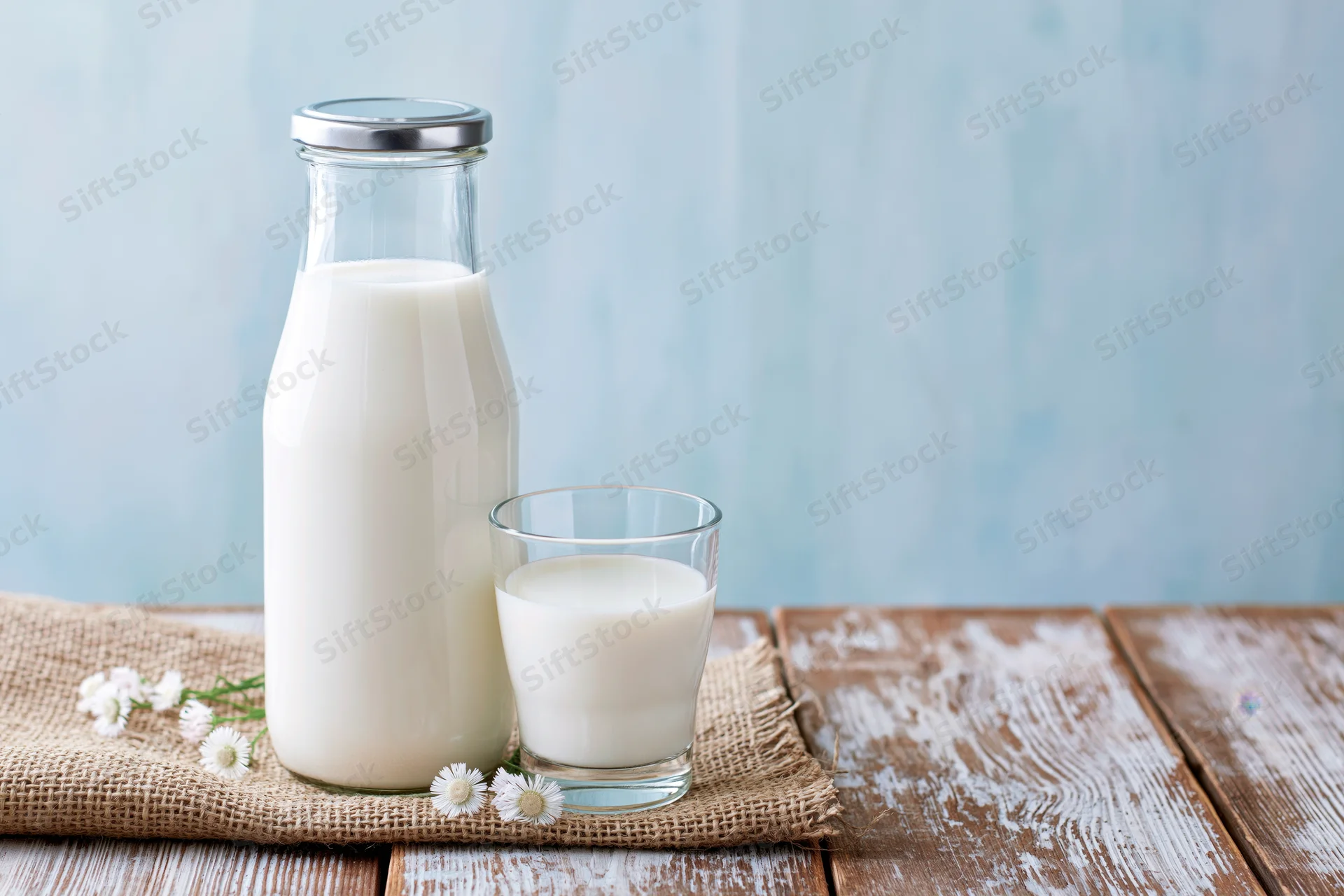 Glass milk bottle with metal lid and a short drinking glass on a wooden table with burlap and small white flowers