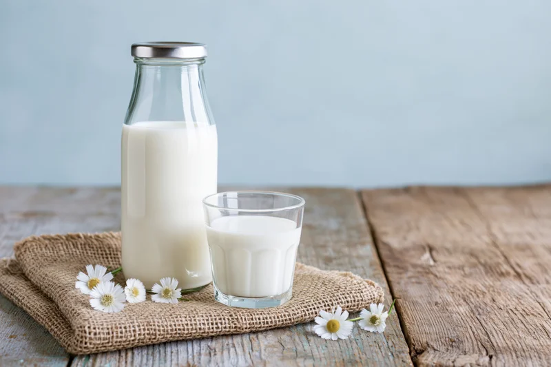 Glass milk bottle and short drinking glass filled with milk on burlap cloth with daisies on wooden table