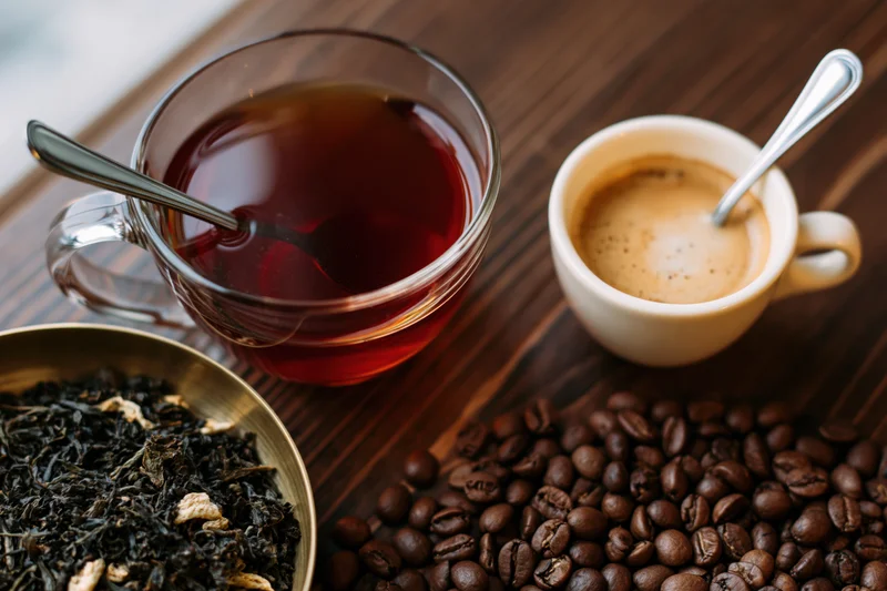 Glass mug of deep red black tea with spoon and ceramic cup of coffee on wooden table beside coffee beans and dried tea leaves