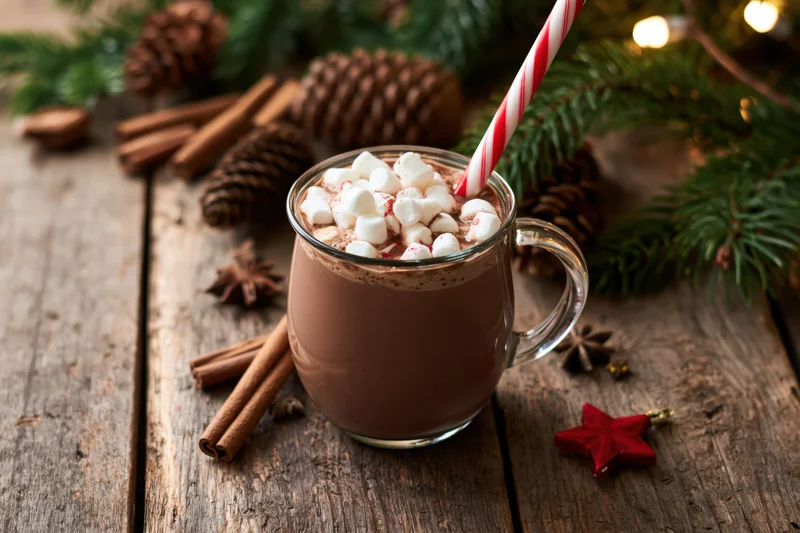 Glass mug of hot chocolate with marshmallows and striped straw on wooden table surrounded by pinecones and holiday decorations
