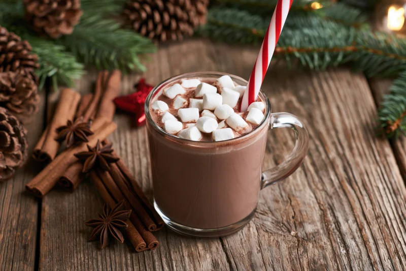 Glass mug of hot chocolate with marshmallows and striped straw on wooden table surrounded by pinecones and cinnamon sticks