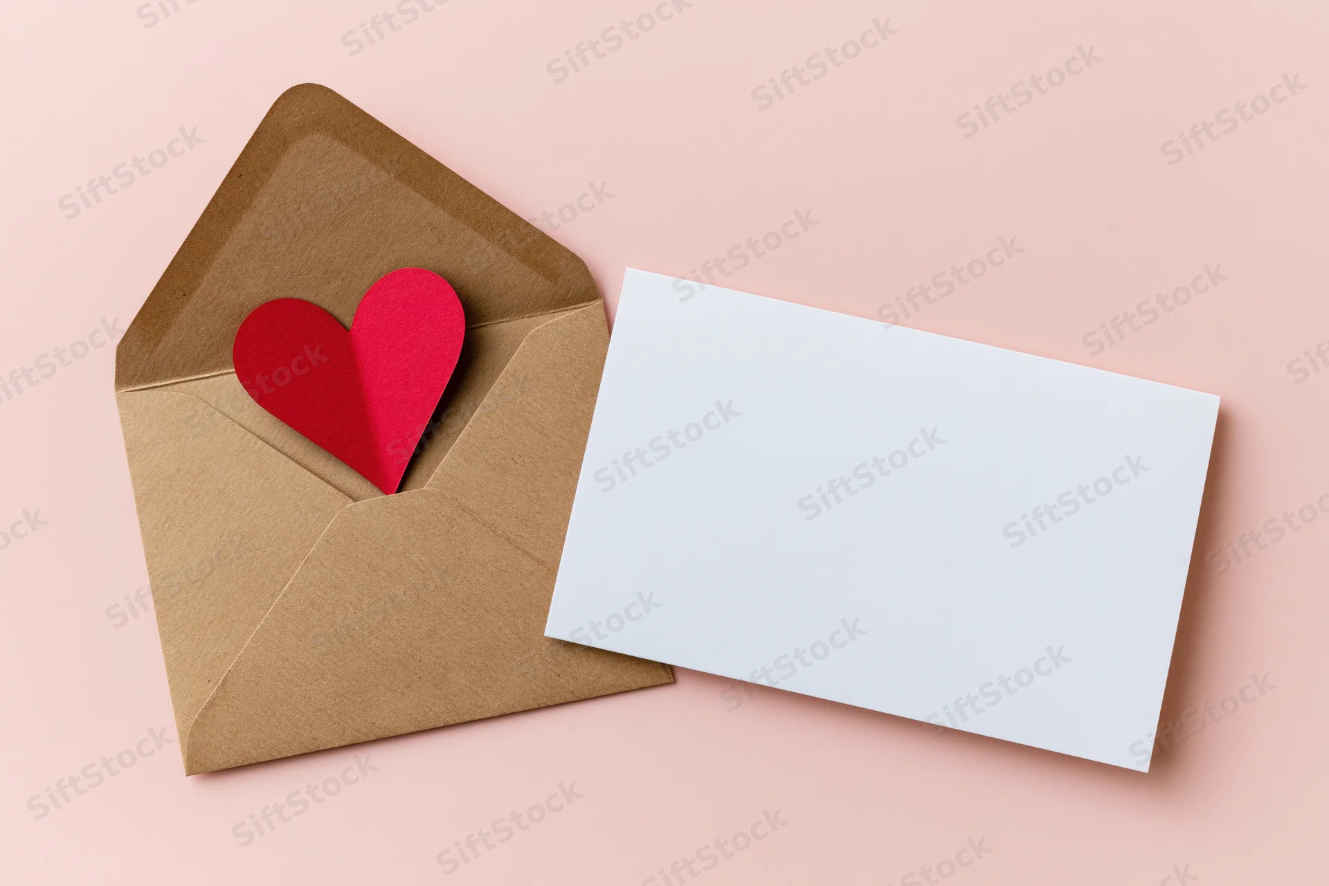 Open kraft envelope with red paper heart peeking out beside blank white card on pink surface