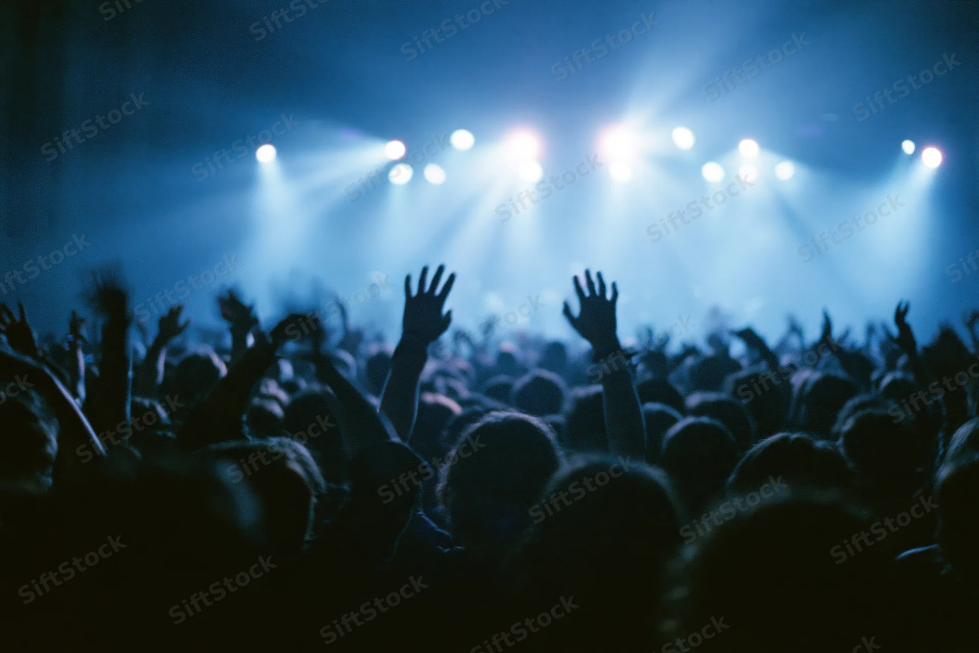 Crowd of concertgoers with hands raised toward bright stage lights at live music event