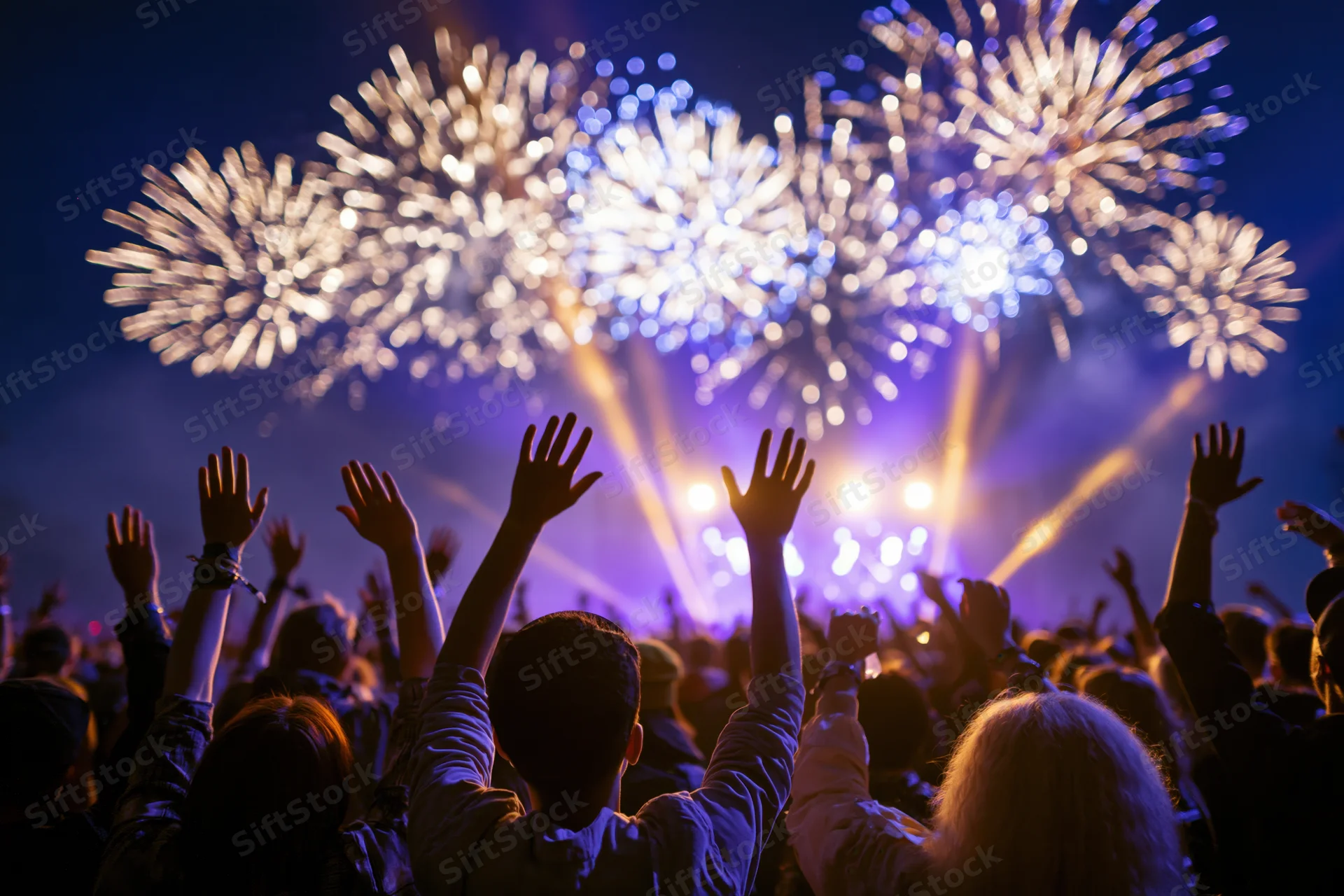 Crowd from behind with arms raised watching fireworks at night outdoors