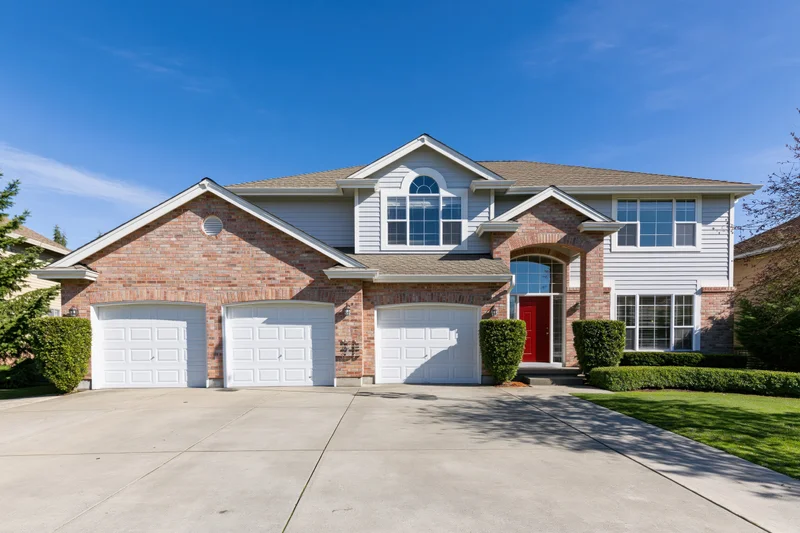 Two-story suburban house with brick and grey siding, red front door, three garages, and concrete driveway under clear blue sky