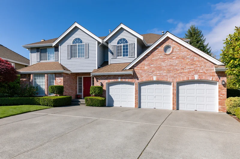 Two-story suburban house with brick and grey siding, red front door, three garages, and landscaped front yard