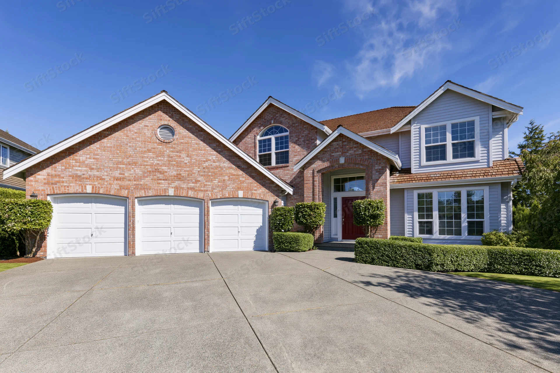 Two-story suburban house with brick and grey siding, white garage doors, and a concrete driveway under a clear blue sky