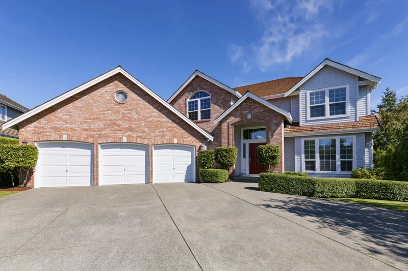 Two-story suburban house with brick and grey siding, white garage doors, and a concrete driveway under a clear blue sky