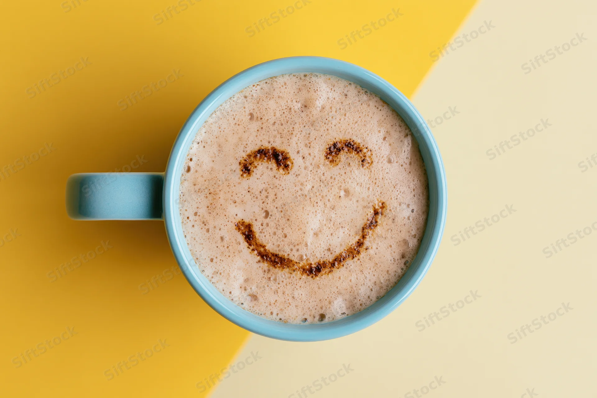 Blue ceramic mug filled with frothy cappuccino featuring a smiley face design on the foam, set on a yellow and beige background
