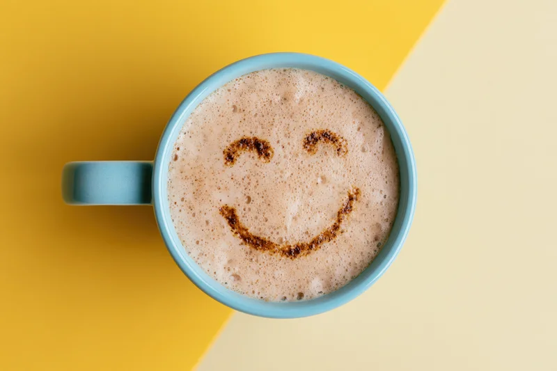 Blue ceramic mug filled with frothy cappuccino featuring a smiley face design on the foam, set on a yellow and beige background