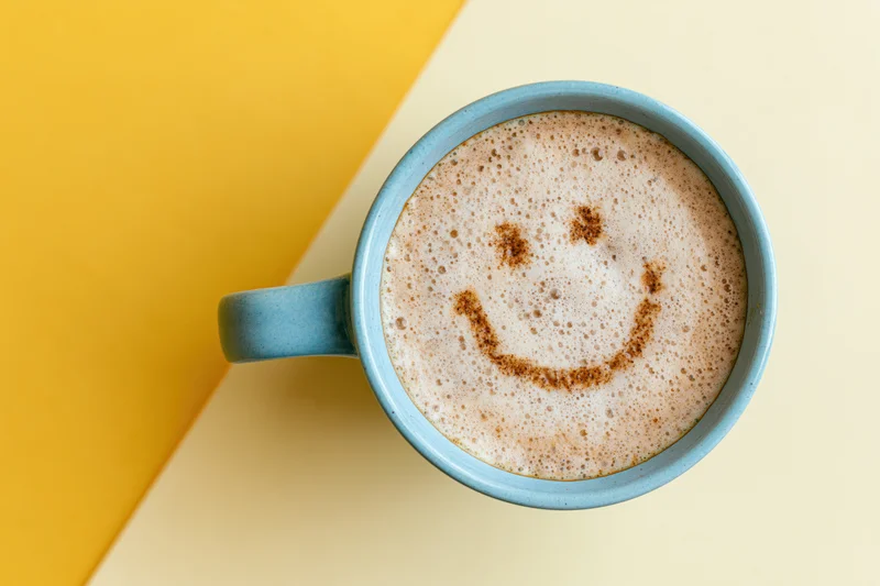 Blue ceramic mug filled with frothy cappuccino featuring a cinnamon smiley face on a yellow and beige surface