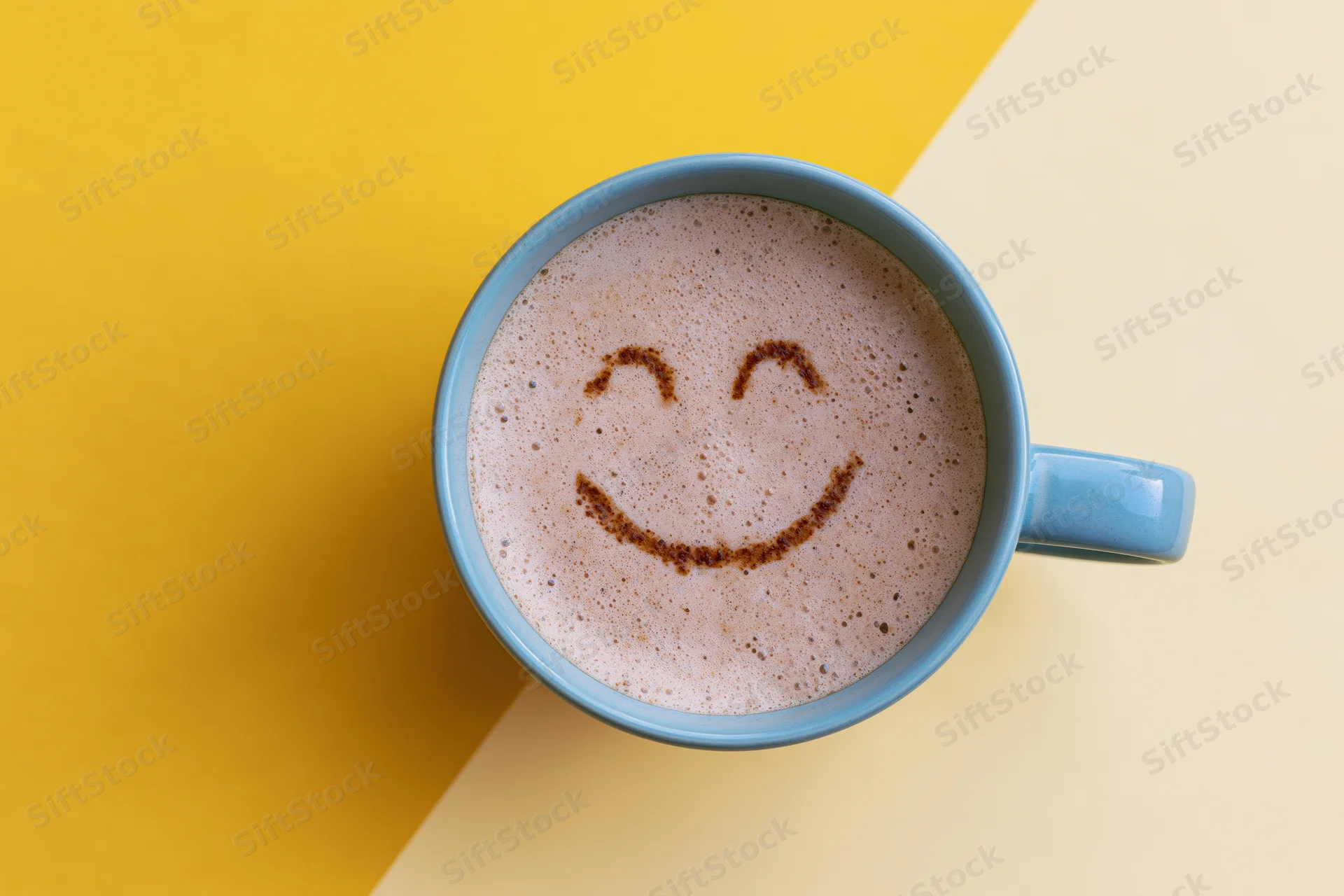 Blue ceramic mug with frothy cappuccino featuring a smiley face drawn in foam on a yellow and cream background