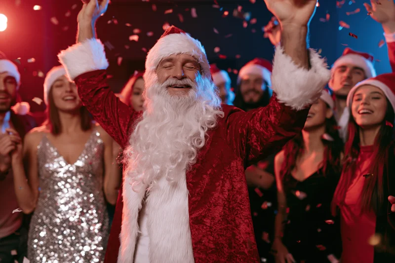 Santa dancing with arms raised at a festive party surrounded by smiling people in holiday attire