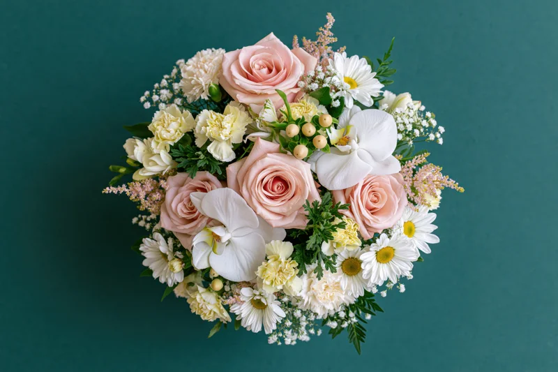Overhead view of a mixed flower bouquet with pink roses, white orchids, daisies and greenery centered on a teal background