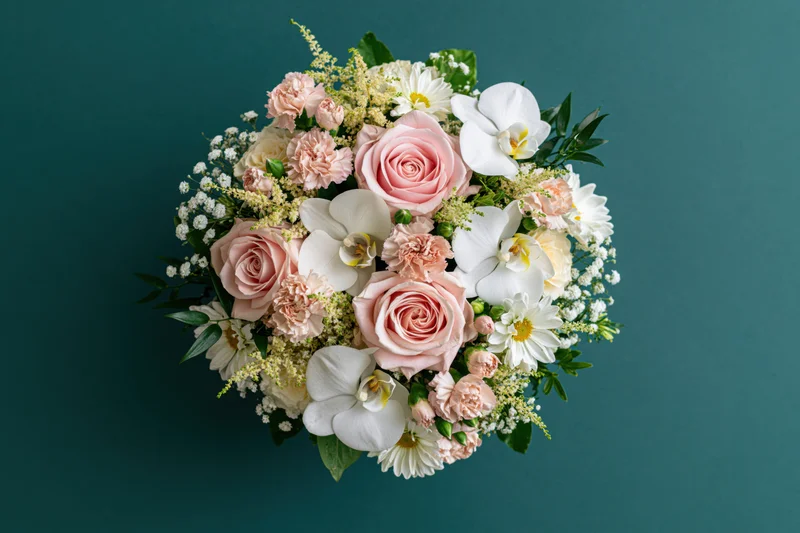 Overhead view of a round mixed flower bouquet with pink roses, white orchids, and greenery on a teal background