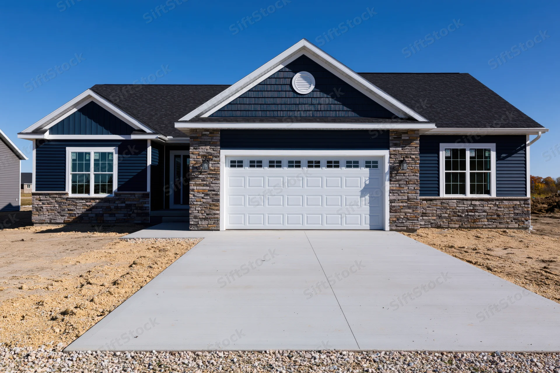 Newly constructed single-story ranch house with navy blue siding and stone accents, featuring a white garage door and concrete driveway under a clear blue sky