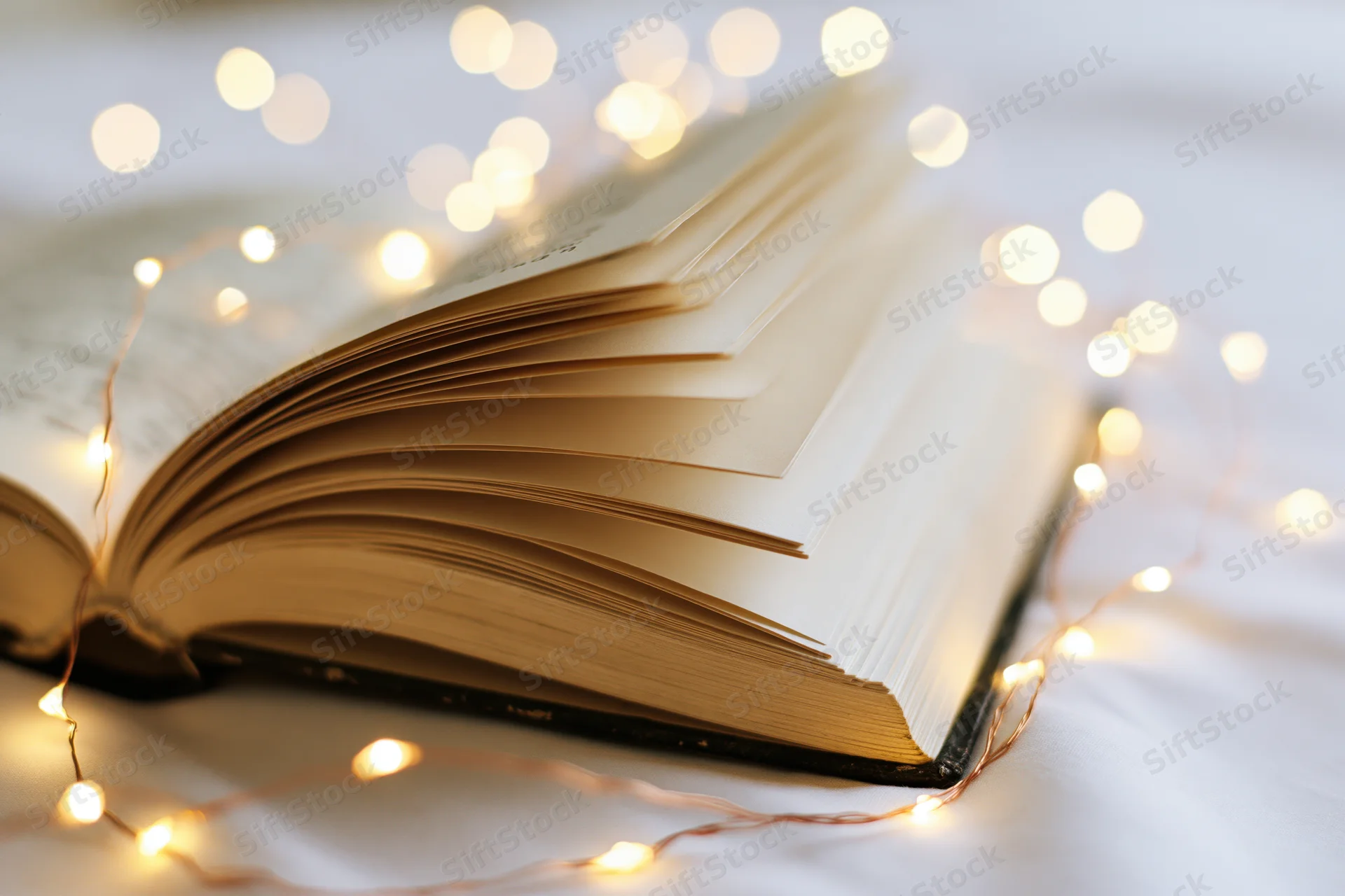 Open hardcover book with fanned pages and string lights glowing softly on white surface