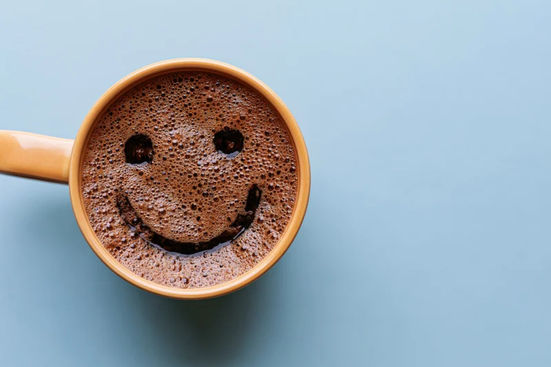 Overhead view of a ceramic coffee mug with smiley face foam on blue background