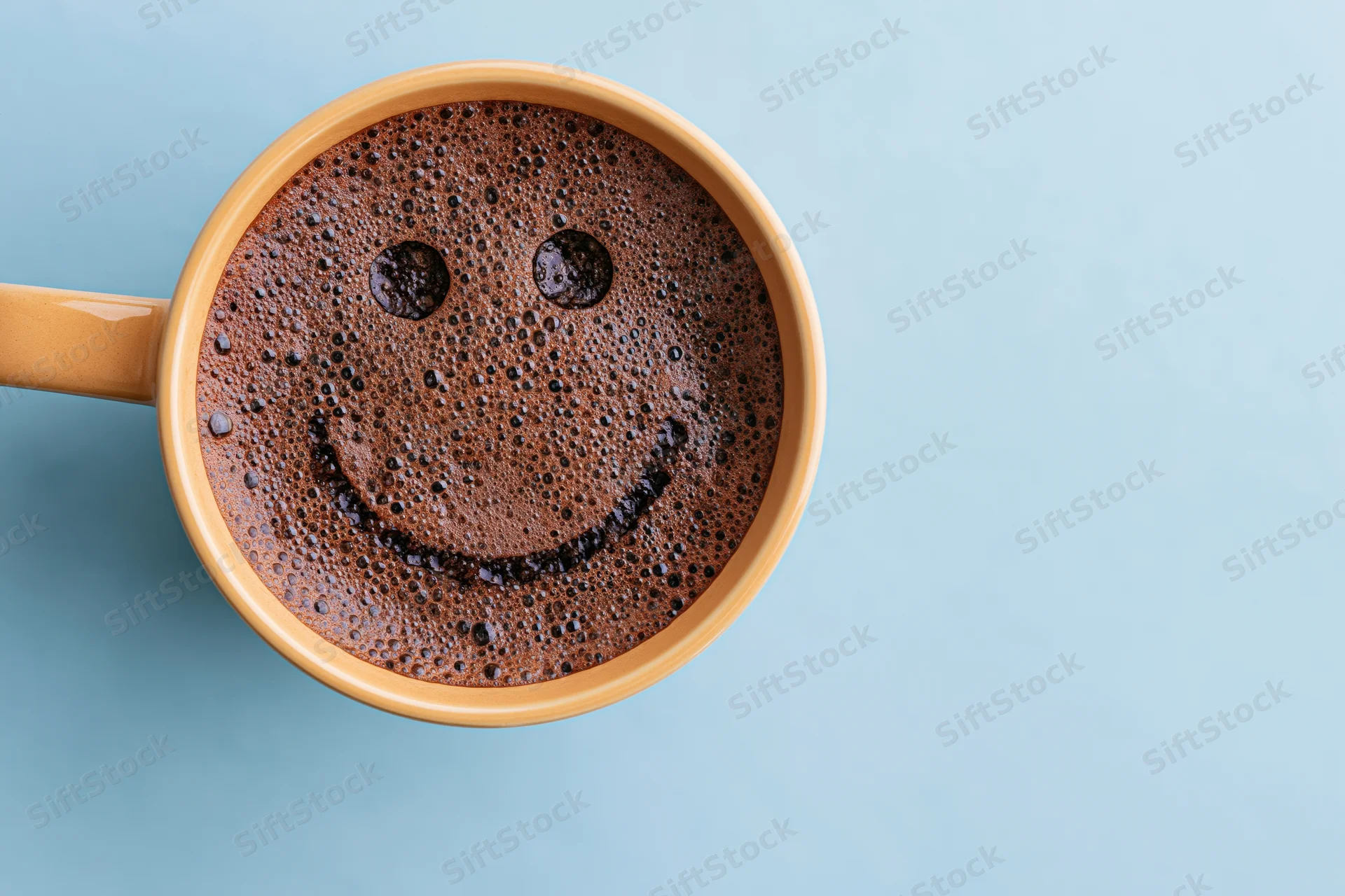 Overhead view of a ceramic coffee mug with smiley face foam on blue background