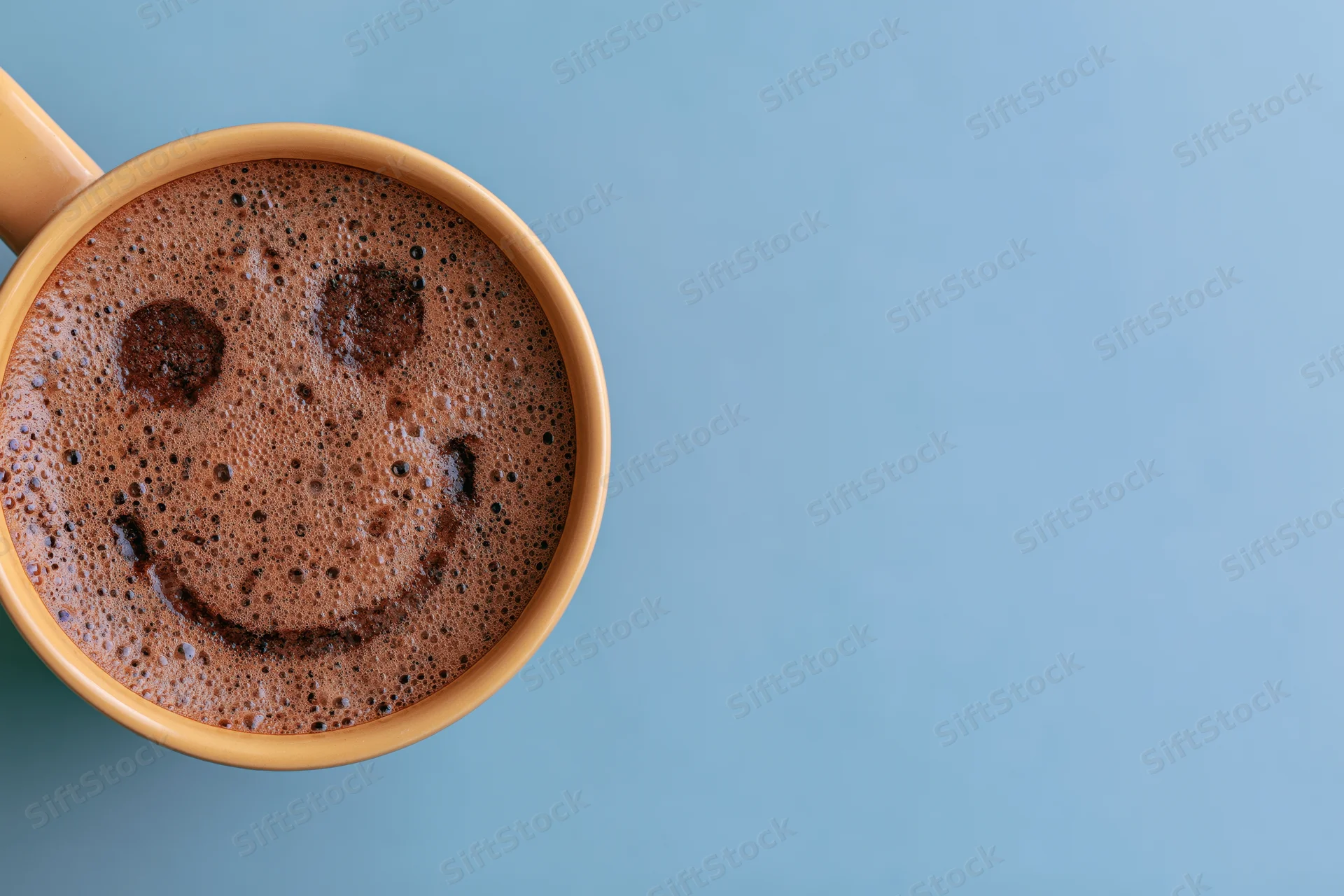 Overhead close-up of a ceramic coffee mug filled with foamy coffee showing a smiley face on a blue background with copyspace