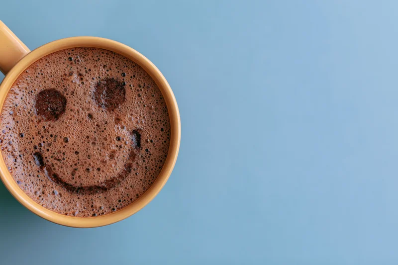 Overhead close-up of a ceramic coffee mug filled with foamy coffee showing a smiley face on a blue background with copyspace