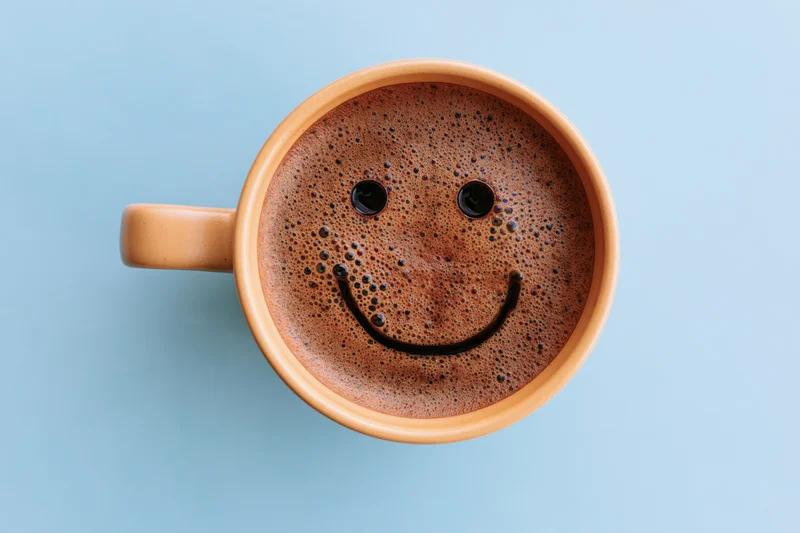 Overhead view of a ceramic coffee mug with smiley face drawn in the foam on a blue background
