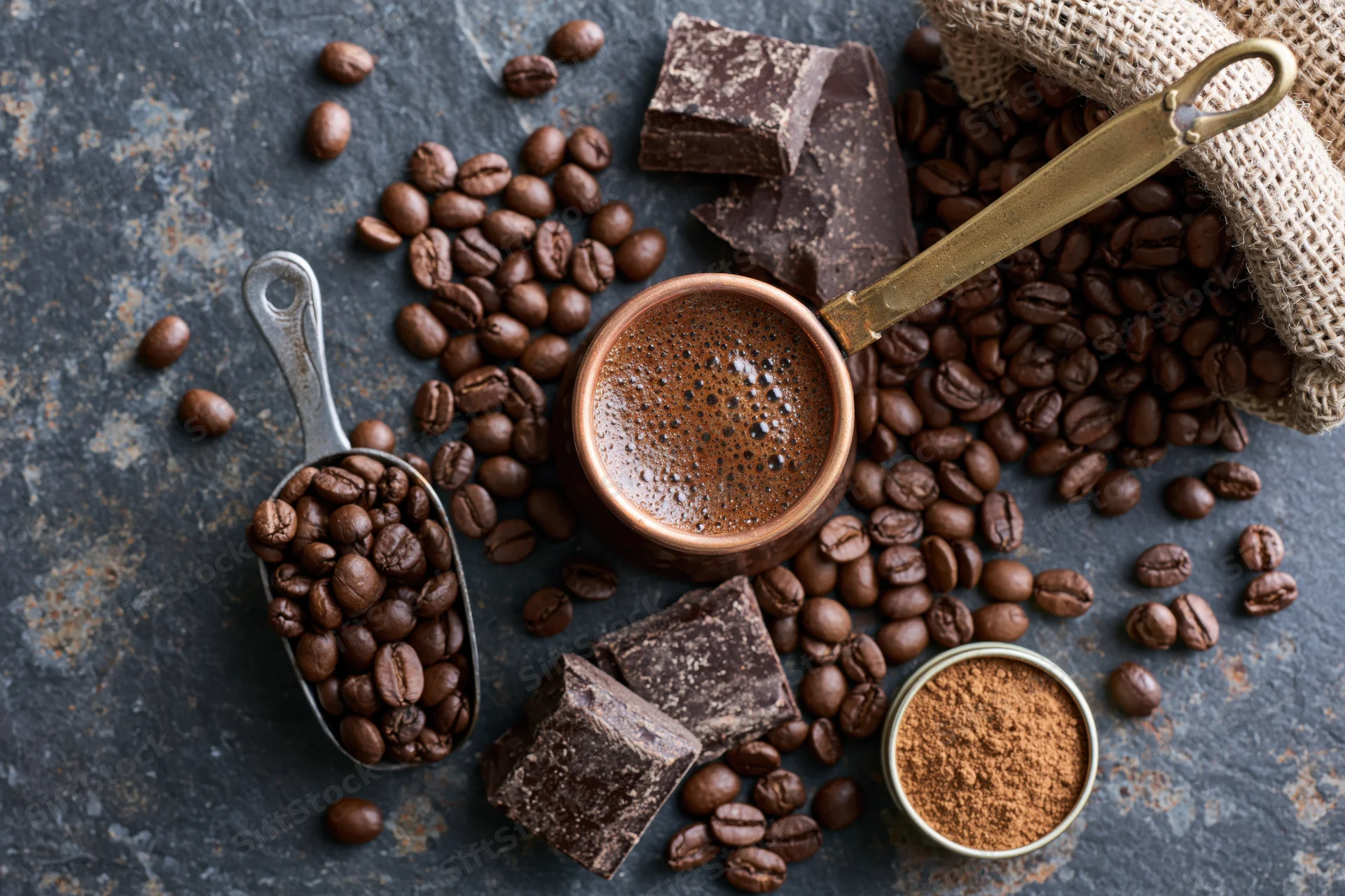 Copper cezve filled with frothy coffee surrounded by beans, chocolate, and ground coffee on dark rustic surface