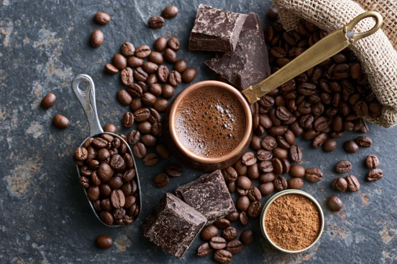 Copper cezve filled with frothy coffee surrounded by beans, chocolate, and ground coffee on dark rustic surface