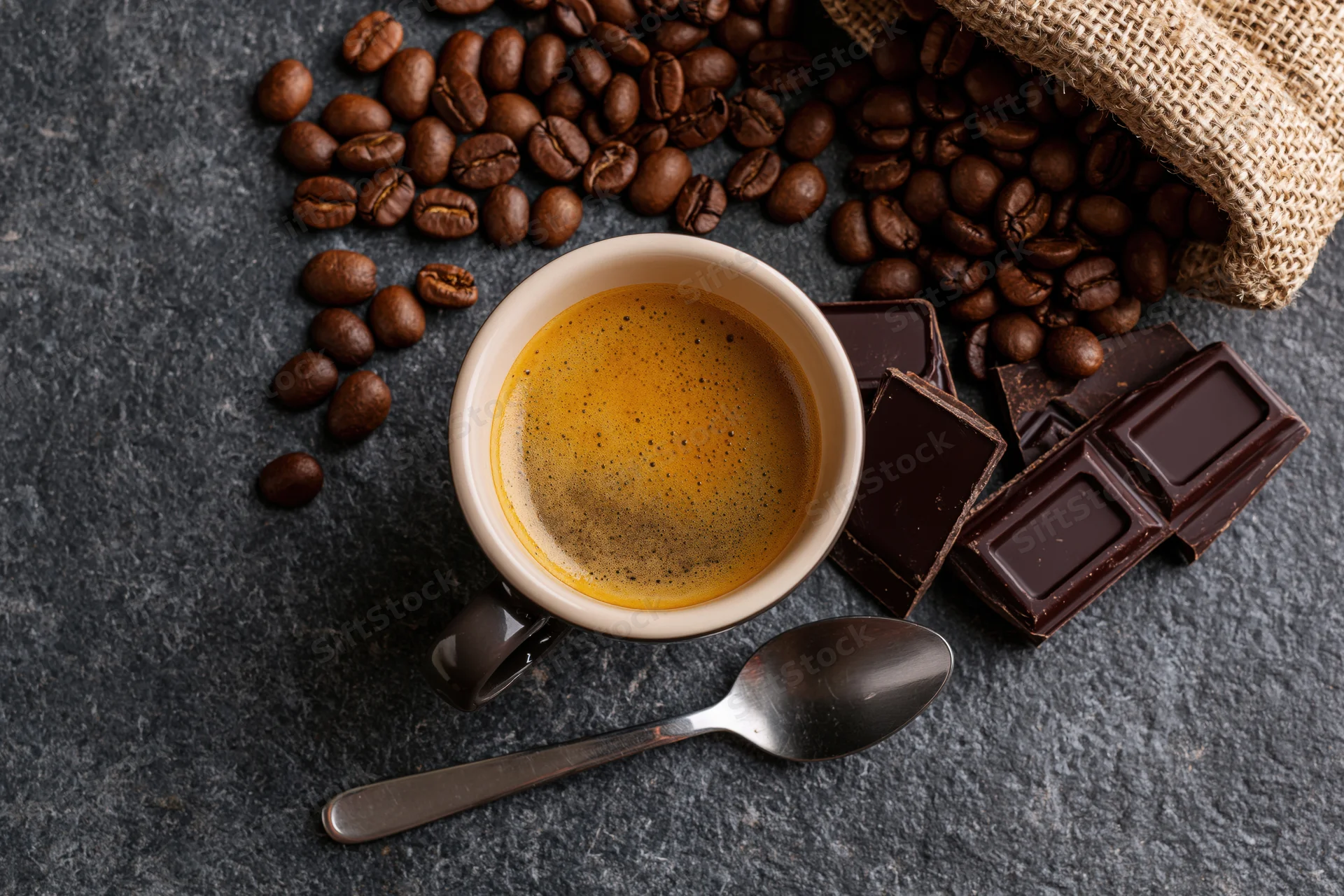 Overhead flat lay of a ceramic espresso cup filled with coffee and crema, surrounded by coffee beans, chocolate pieces, and a spoon on a dark stone surface