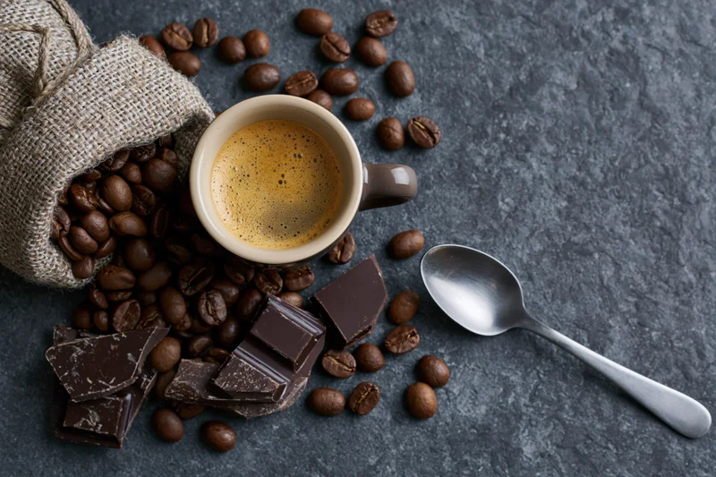Overhead flat lay of ceramic espresso cup with frothy coffee, scattered beans, chocolate pieces, spoon, and burlap bag on dark stone surface