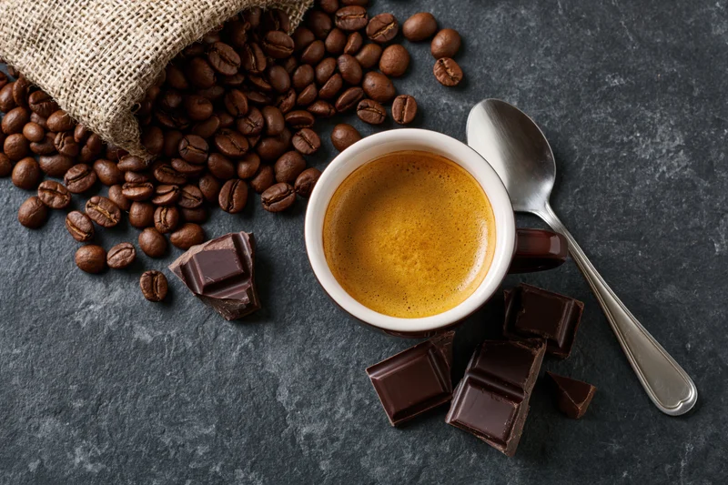 Overhead flat lay of a ceramic espresso cup filled with golden crema surrounded by roasted coffee beans, dark chocolate pieces, a spoon, and a burlap bag on a slate surface