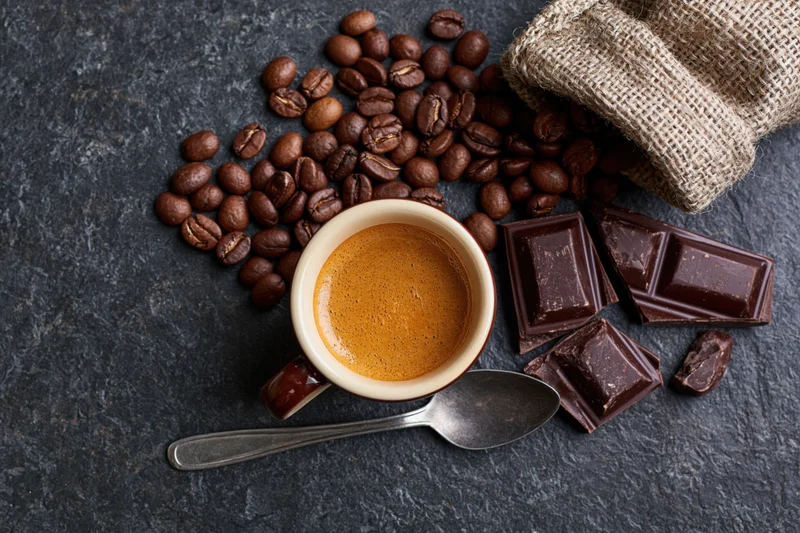 Overhead flat lay of a ceramic espresso cup filled with golden crema surrounded by roasted coffee beans, dark chocolate pieces, and a burlap sack on a slate surface with a metal spoon beside it