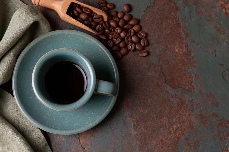 Overhead flat lay of a teal ceramic coffee cup with saucer, coffee beans and wooden scoop on a rustic metal surface with linen cloth