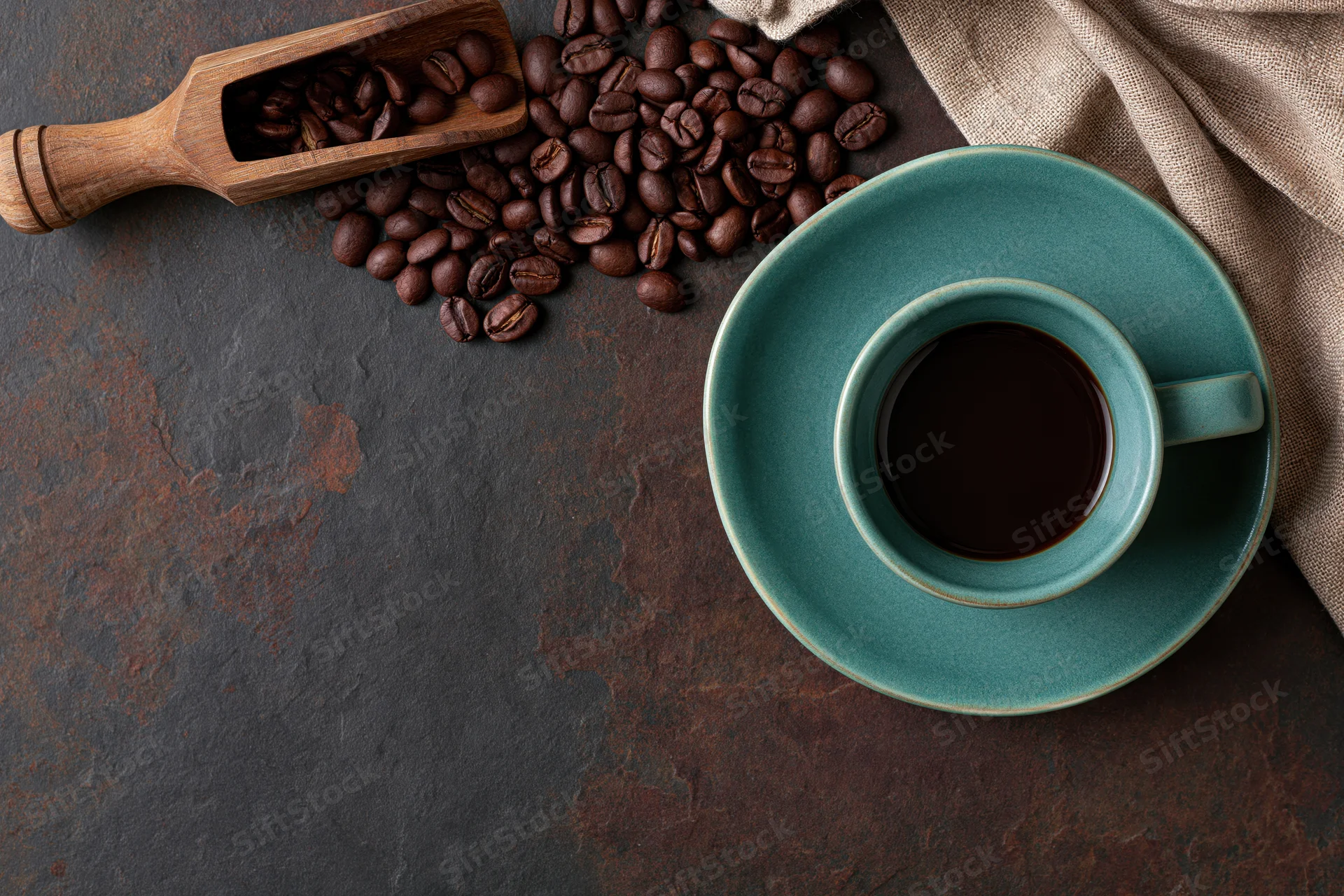 Teal ceramic coffee cup filled with black coffee on dark textured surface beside roasted beans and wooden scoop