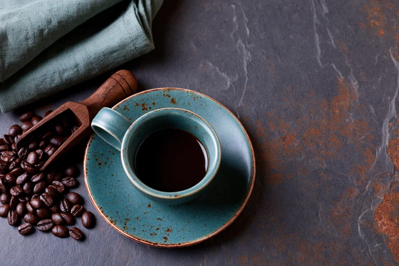 Teal ceramic coffee cup filled with dark coffee on a marble countertop beside coffee beans and a wooden scoop