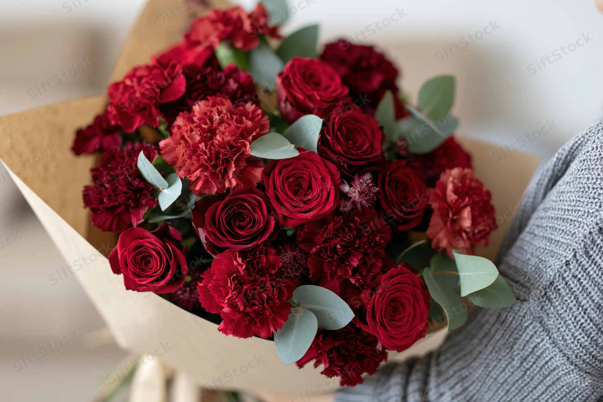 Person holding a large mixed bouquet of deep red and crimson roses and carnations wrapped in kraft paper