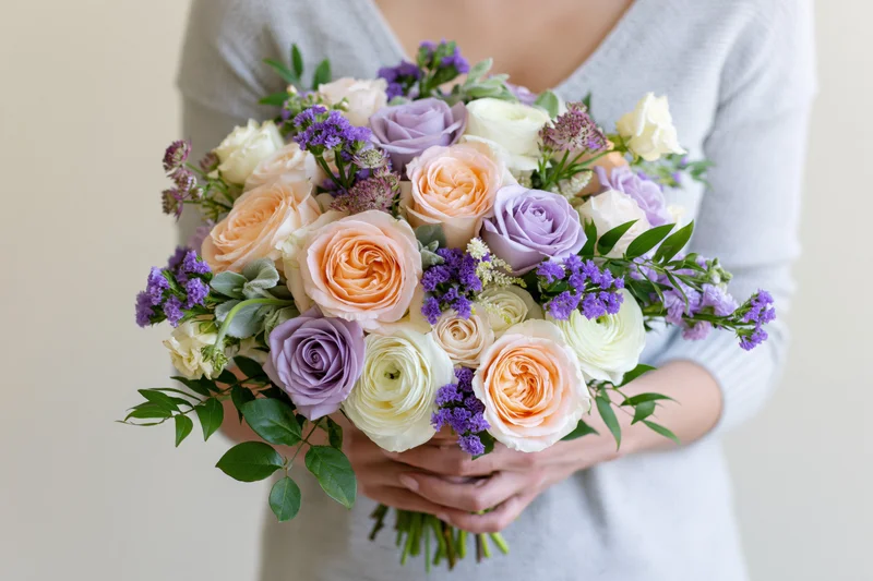 Woman holding a lush mixed bouquet of peach, lavender, and white roses with greenery