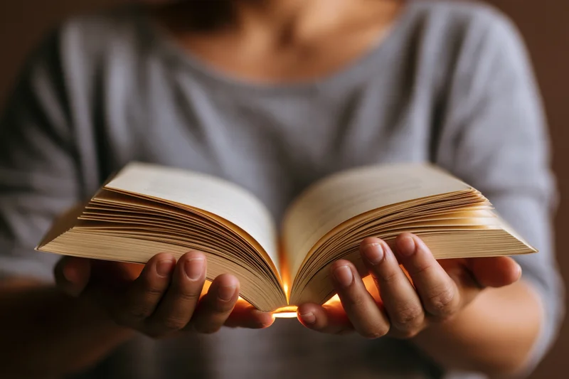 Woman holding open book with glowing pages, hands visible, soft warm light between pages