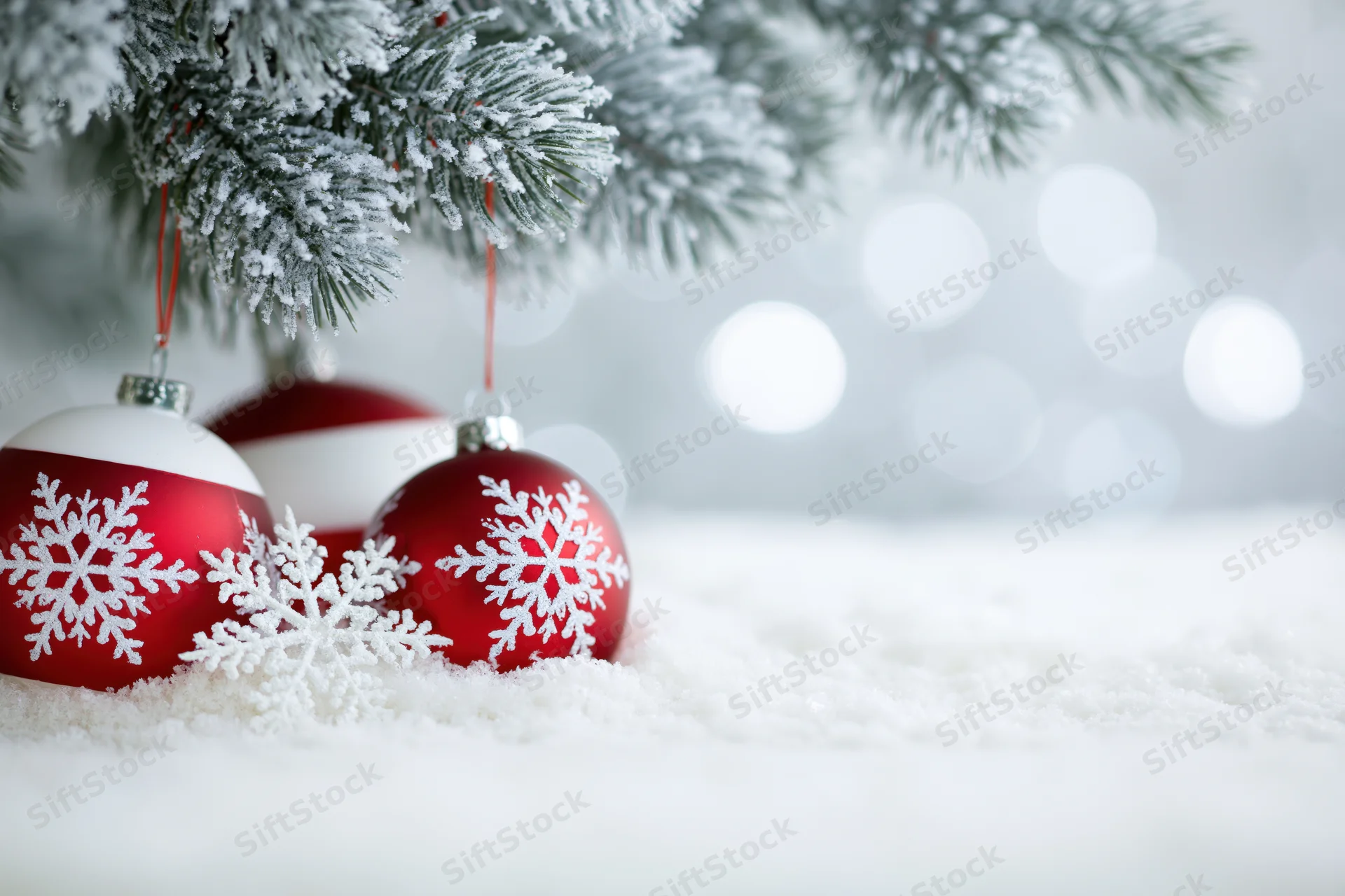 Red and white snowflake ornaments resting under a snowy pine branch with bokeh lights in the background