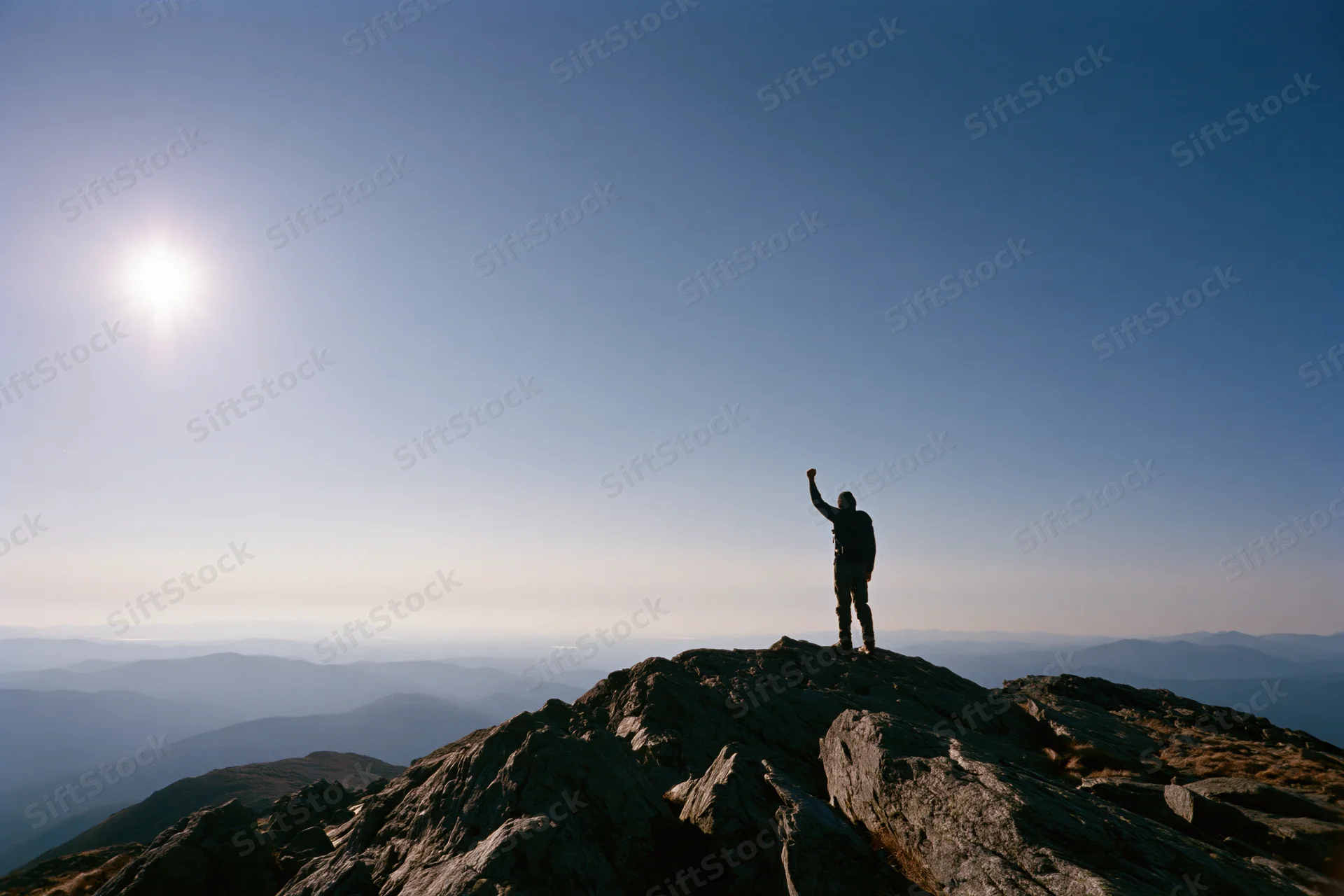 Hiker standing on rocky mountain summit with raised arm under bright sun