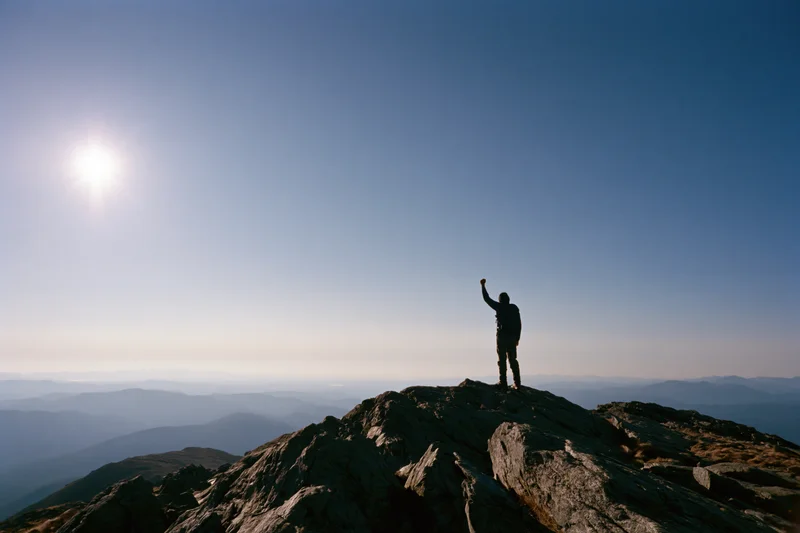 Hiker standing on rocky mountain summit with raised arm under bright sun