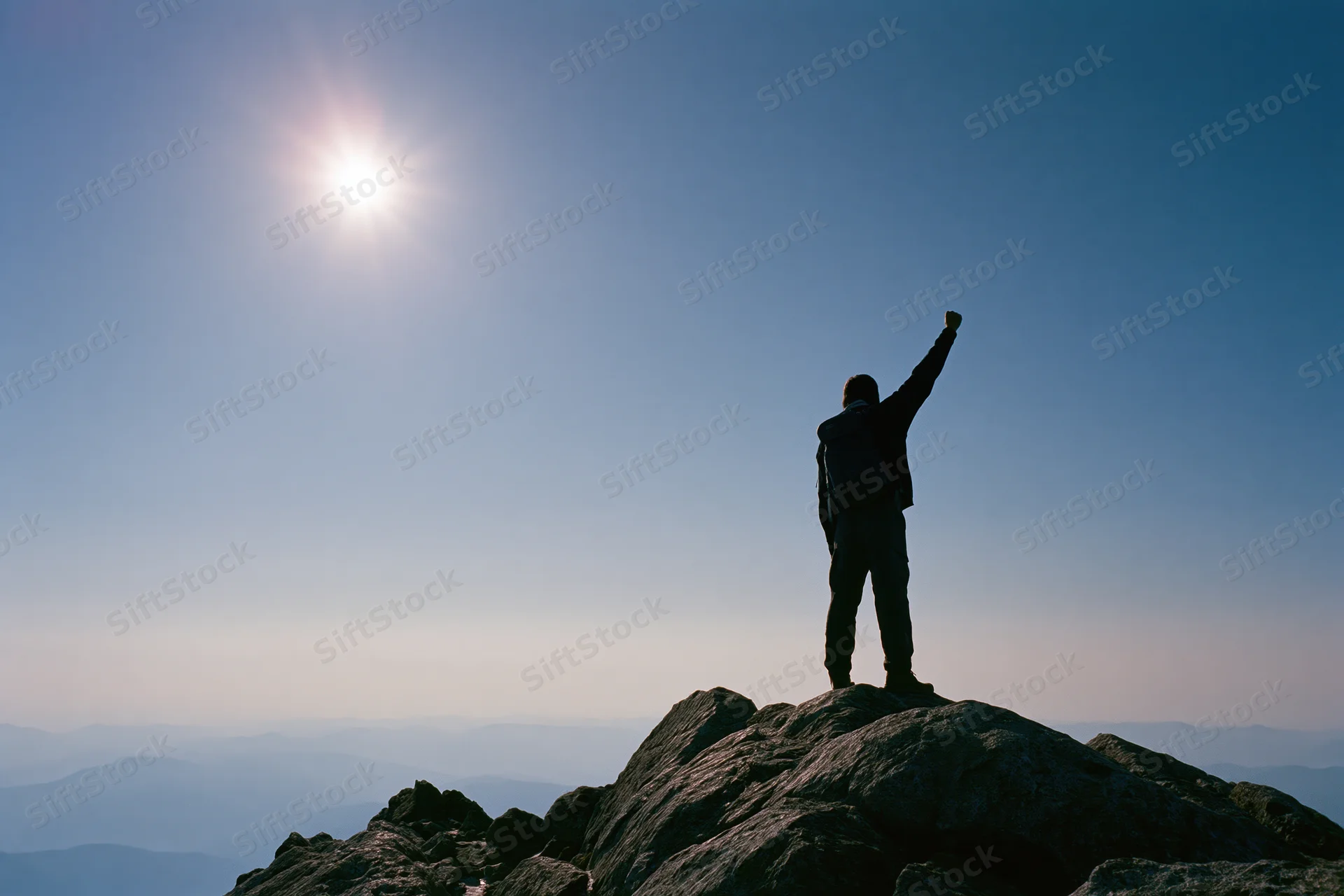 Hiker standing on rocky mountain summit with arm raised toward sun