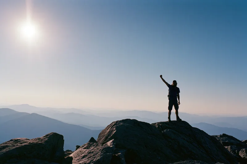 Hiker standing on rocky mountain summit with arm raised against sunrise sky