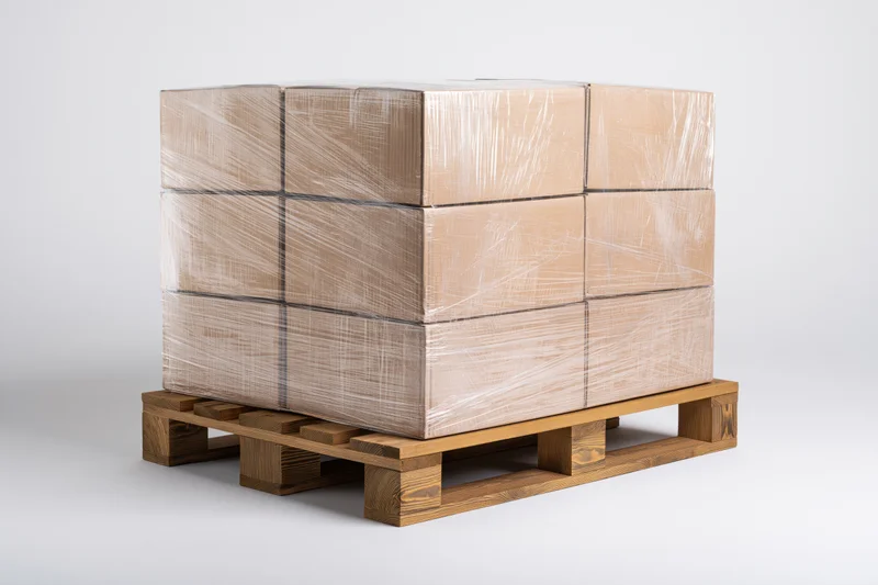 Stack of cardboard boxes wrapped in clear stretch plastic on wooden pallet against white background